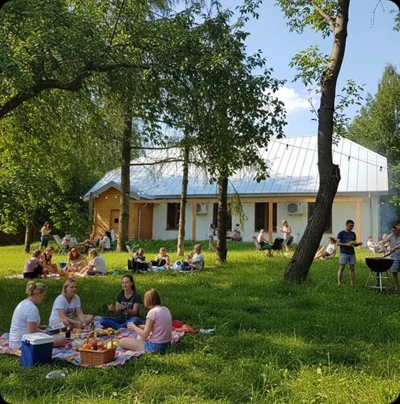 grupa ludzi na kocu uczestniczy w pikniku, w tle natura, drzewa, drewniany dom